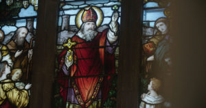 A stained glass representation of St. Patrick holding a cross in his left hand and a clover upheld in his right with crowds of people looking towards him.